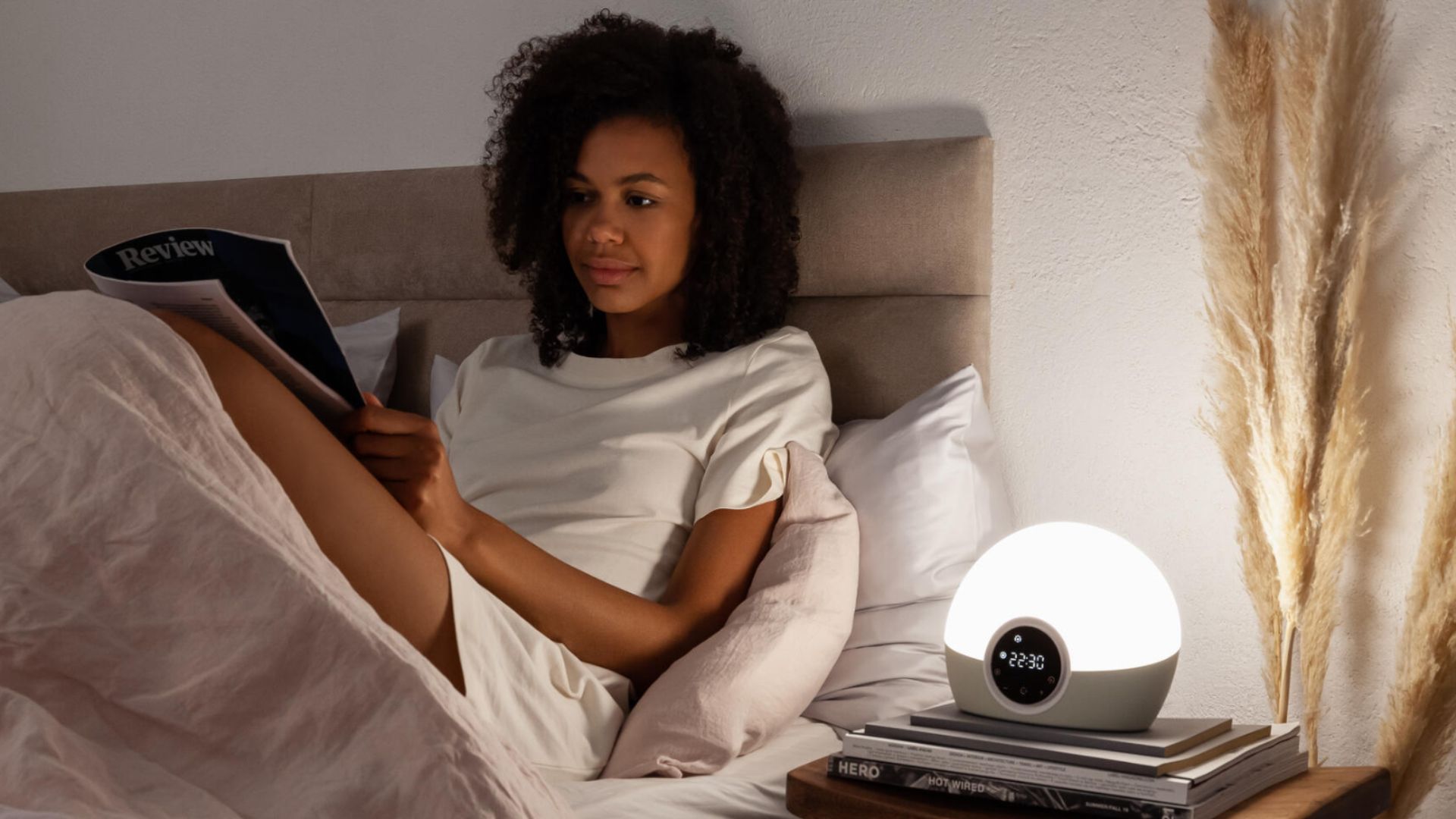 A woman in bed reads next to Lumie's alarm as the sun rises over a pile of books on a bedside table and pampus grass.