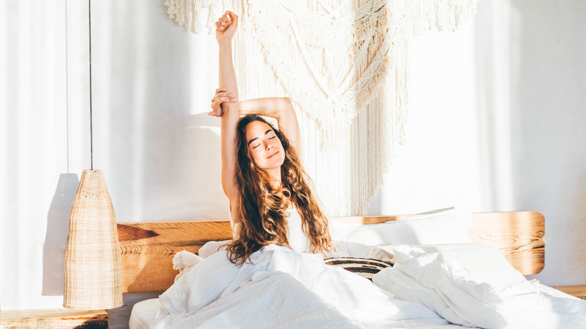 A woman with long black hair is sitting on the bed with her arms outstretched in the air as sunlight streams in through her open curtains.