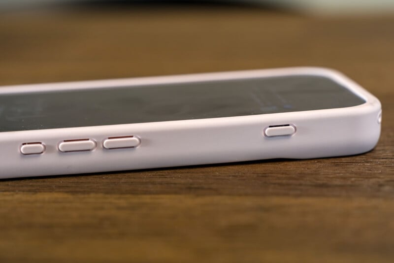 A close-up view of a smartphone with a white or light pink case, showing the volume and power buttons, sitting on a wooden surface.