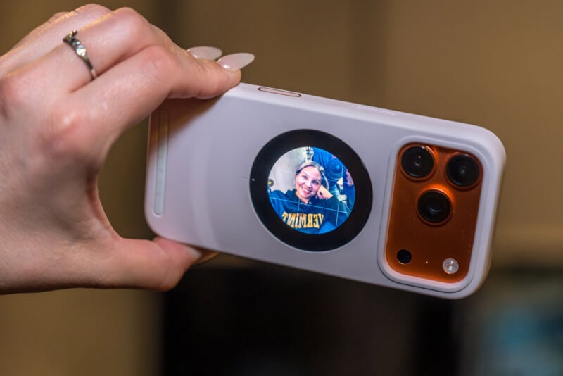 A hand holding a smartphone with a circular display on the back, showing a smiling person taking a selfie. The phone has a white case and three visible camera lenses.