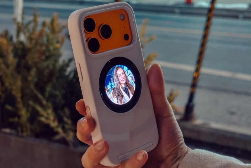 A hand holding a smartphone with a distinctive white case with a small round screen on the back, showing a smiling woman, while large camera lenses appear above the screen. The background shows the street and some greenery.