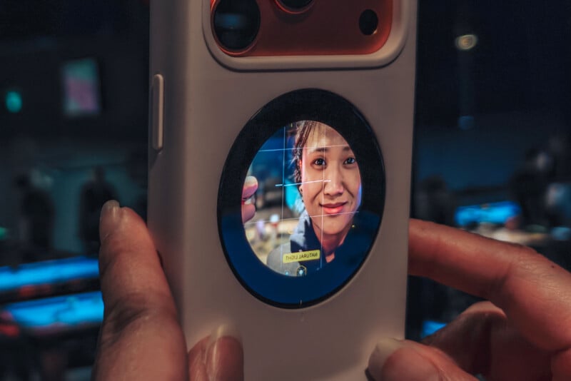 Close-up of a smartphone held in both hands, showing a woman's face smiling into a lens or circular screen in the center of the phone, with a top of hair and a blurred background.