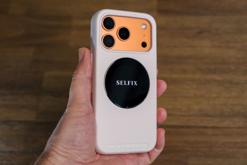 A hand holding a smartphone with a peach colored back, three cameras and a white case. A black circular device with text "DONE ITSELF" attached to the back. The background is a wooden surface.