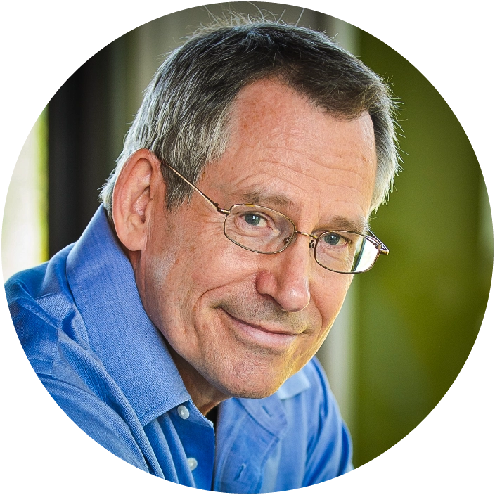 An old man with gray hair and wire-rimmed glasses stands in front of a white board covered with red marker equations and pictures, holding an orange sign in his hands.