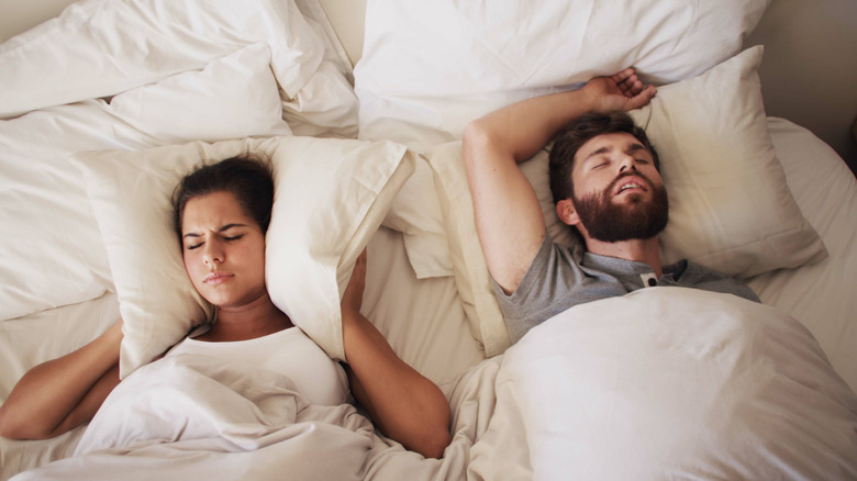 A man and a woman are lying in bed with the women holding a pillow to their ears while the man is snoring