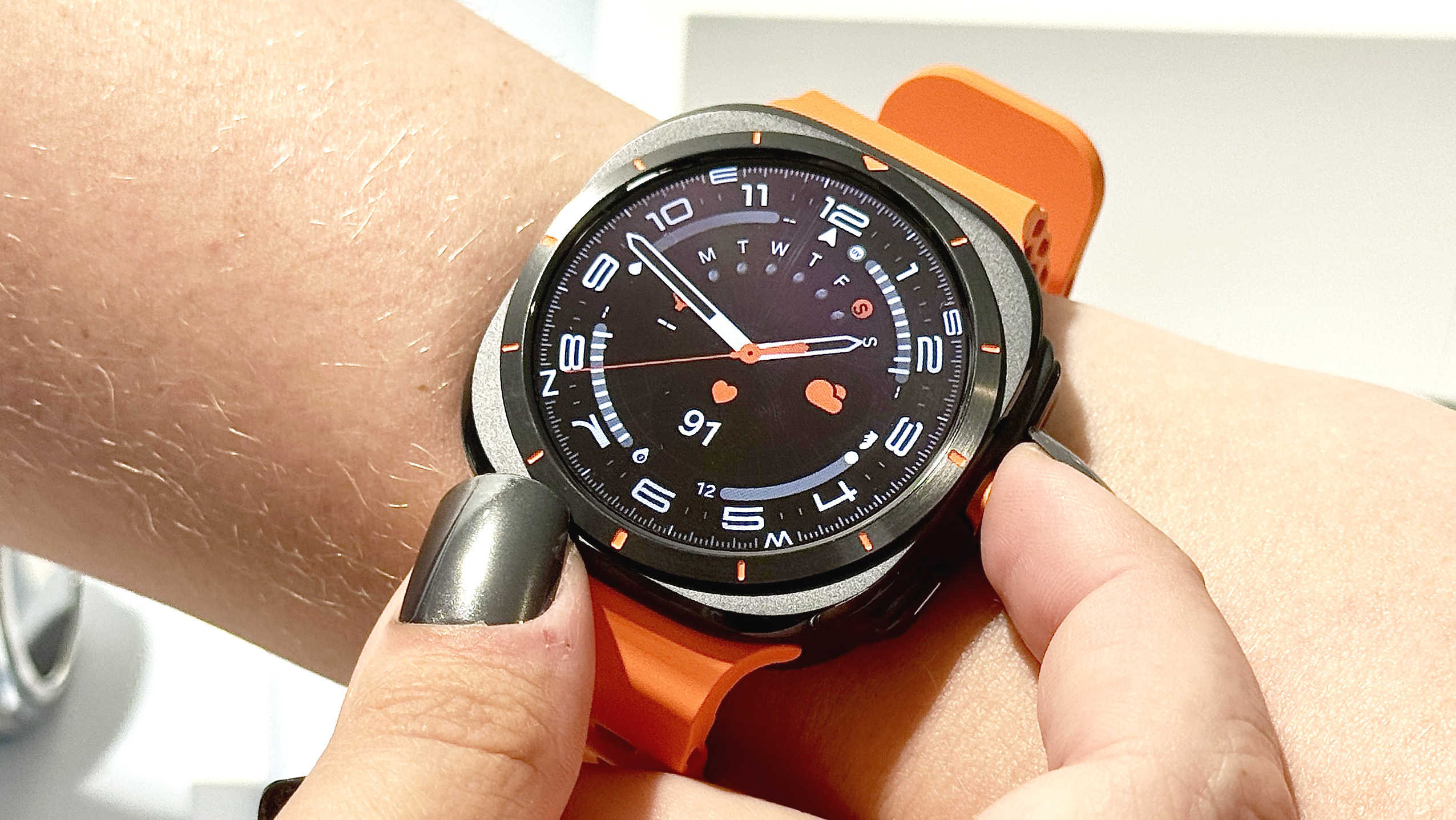 Next to the Samsung Galaxy Watch Ultra in the user's hand with an analog watch face; bright orange cord